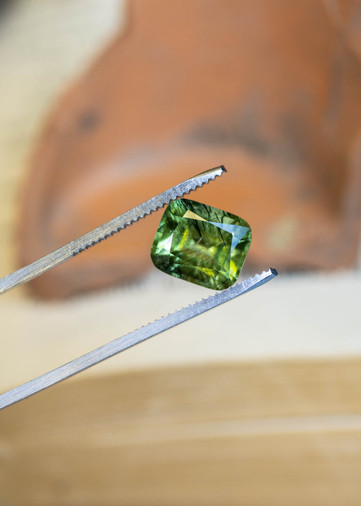 Close-up of a natural Pakistan peridot held in tweezers, showcasing its cushion mix cut, soft sage green color, and intricate crystal inclusions.