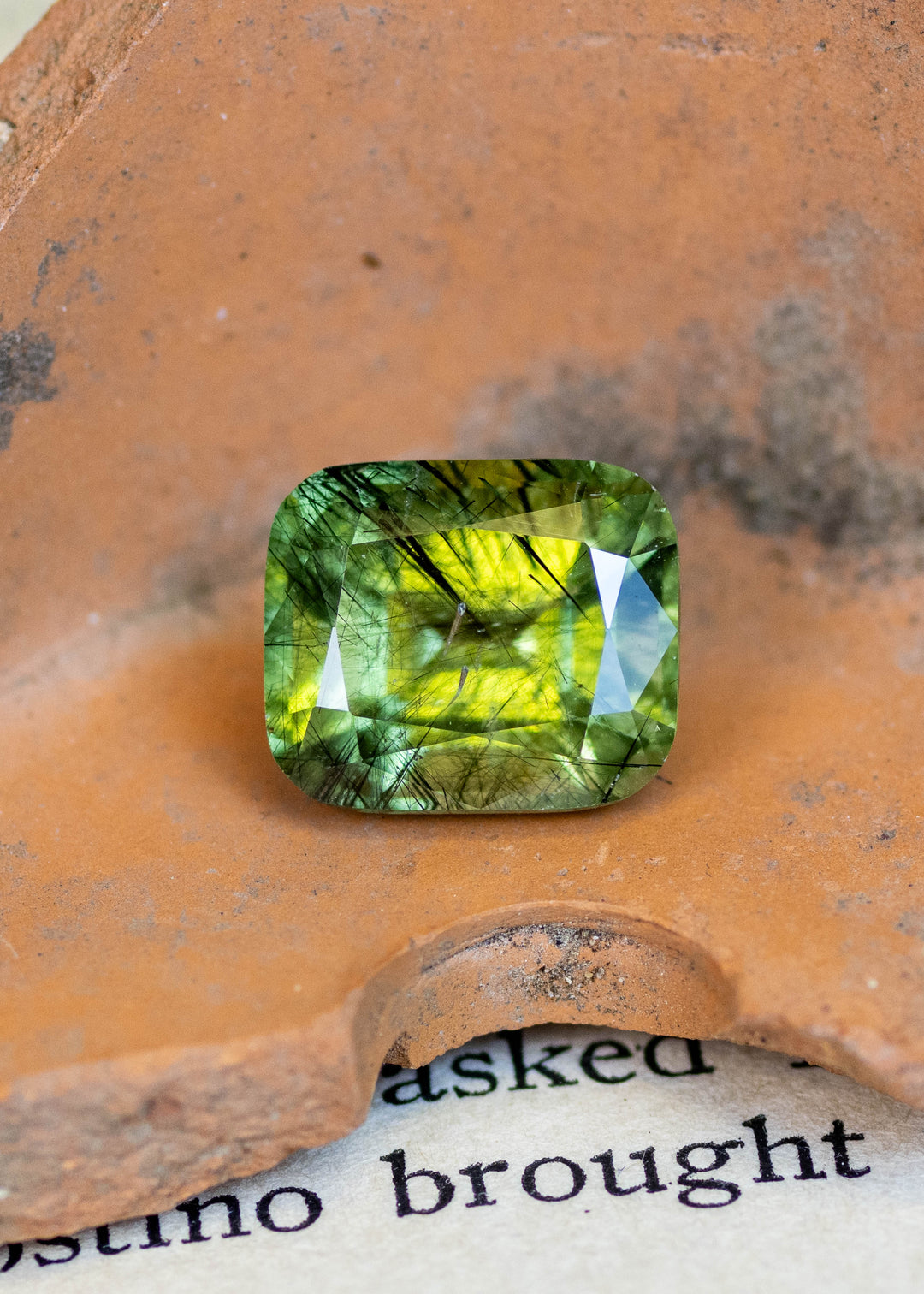 Natural untreated 5.49 carat sage green peridot from Pakistan displayed on terracotta, featuring a cushion mix cut and distinctive natural needle inclusions.