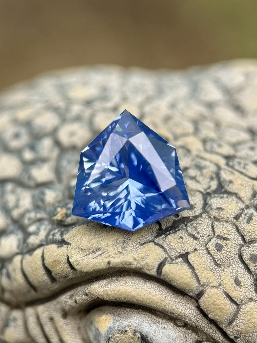 5.10 carat vibrant bright blue sapphire displayed on a textured stone surface, showing intense brilliance and reflective depth of the precision custom trillion shield mix cut, untreated gemstone from Sakaraha, Madagascar.