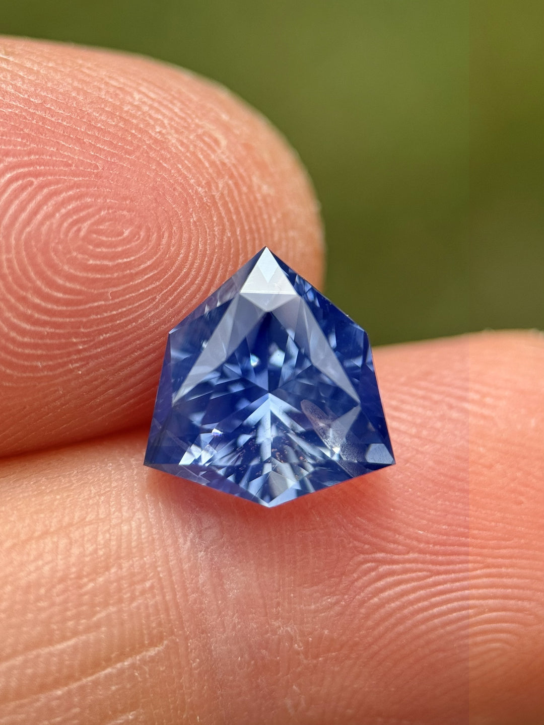 5.10 carat vibrant blue sapphire resting on fingertips under natural light, capturing the gemstone’s radiant luster and intricate reflections of the precision custom trillion shield mix cut, untreated sapphire from Sakaraha, Madagascar.