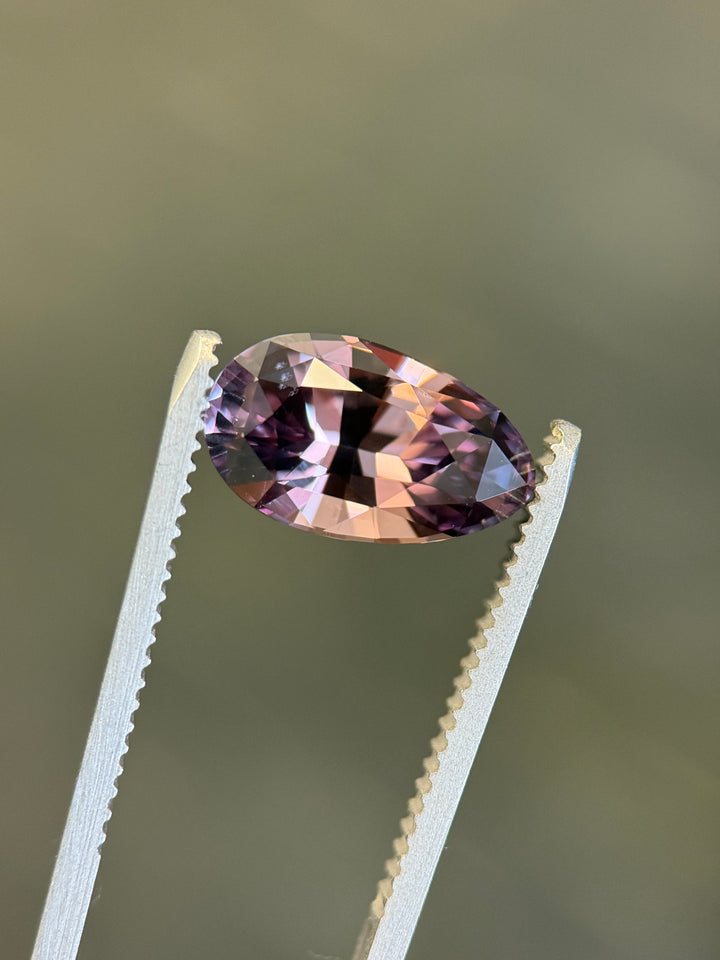 Loose heated sapphire held in tweezers showing the modified brilliant pear cut and shifting purplish red to peach champagne color.