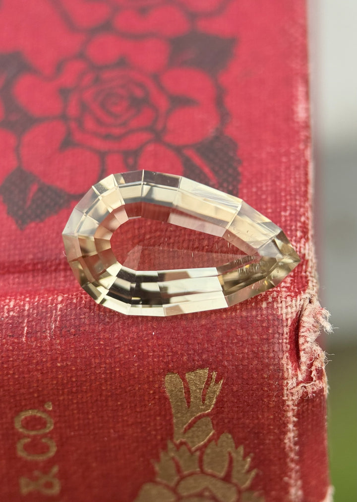 5.36 carat champagne brown tourmaline resting on a red textured book cover, showing custom geometric pear step cut with fine brilliance and elongated shape, untreated gemstone from Congo, Africa.