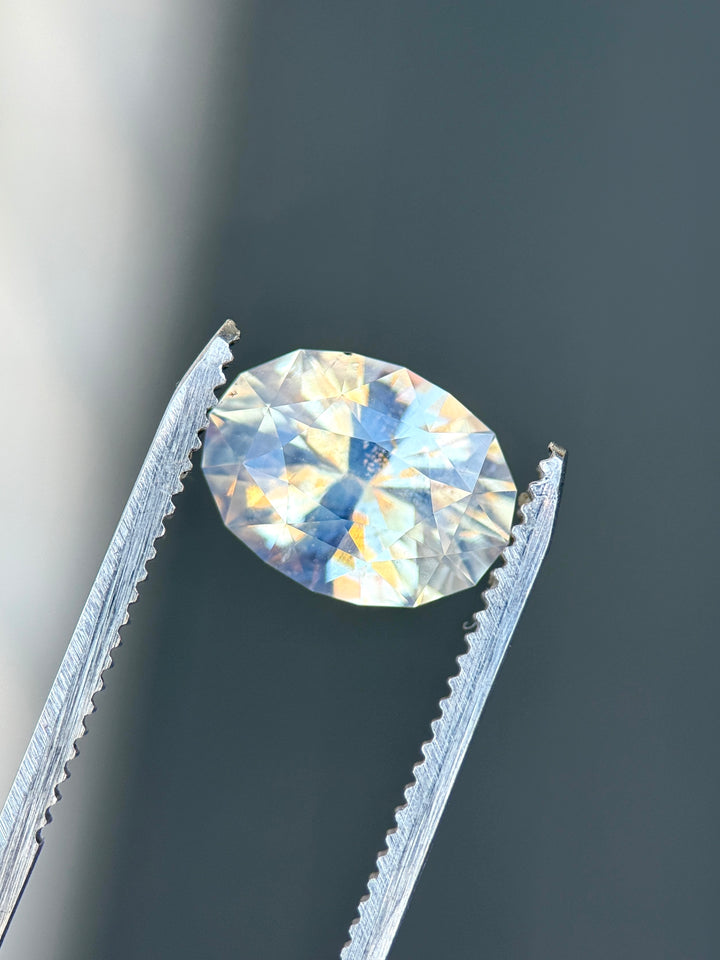 5.56 carat seafoam sapphire held in tweezers highlighting oval brilliant cut and opalescent character.