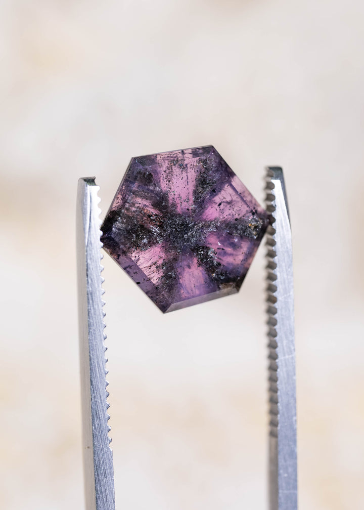 Close-up of a 5.56ct Afghan sapphire held between jeweler's tweezers. Soft lighting reveals its violet-pink coloration, black radial inclusions, and geometric hexagonal portrait cut.