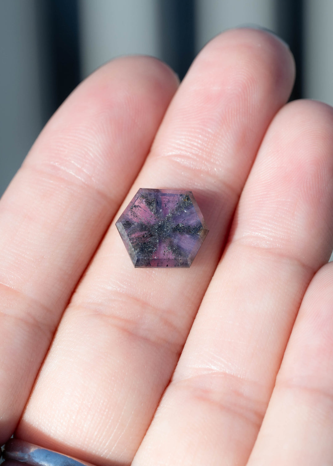 A 5.56ct untreated pink purple sapphire from Afghanistan displayed on fingers for scale. Its broad hexagonal portrait cut showcases the dramatic natural inclusion pattern across a nearly 13 mm face-up silhouette.