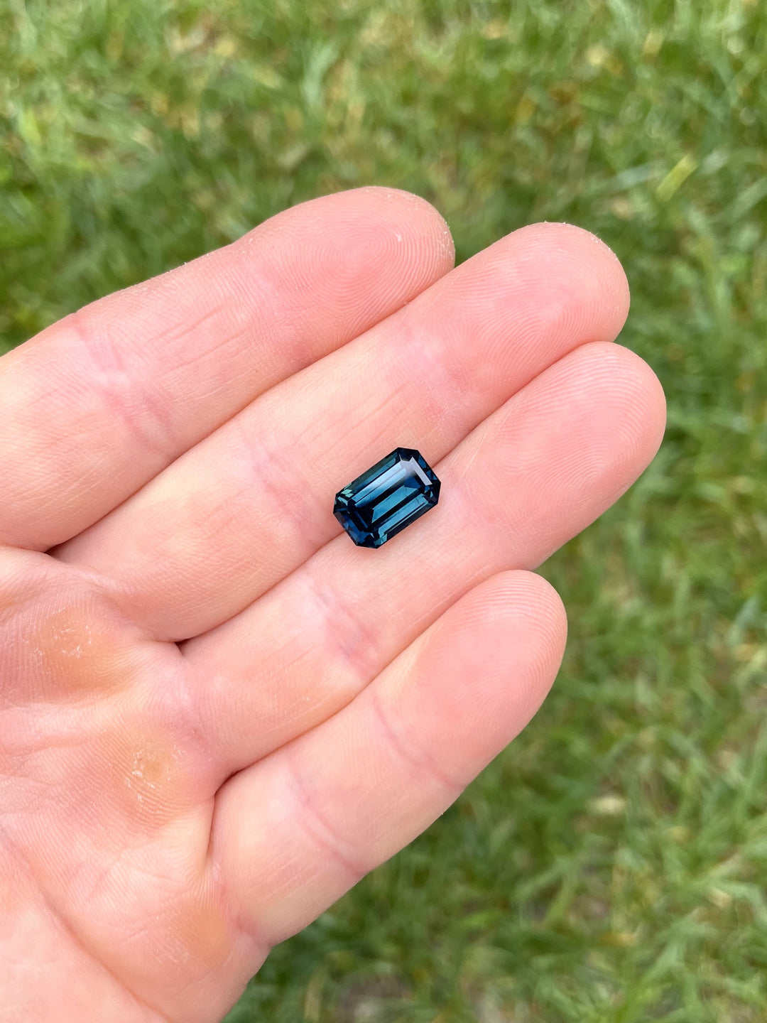 Emerald cut 5.85 carat teal and blue sapphire resting on a hand, showing deep peacock hues and step-cut brilliance from Gombe, Nigeria.