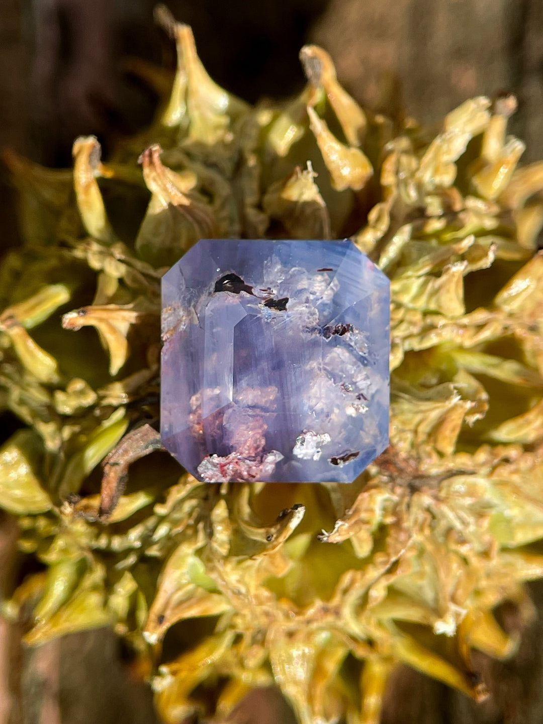 A natural 5.92-carat emerald cut sapphire, blending gray, purple, and brown tones, resting on a textured surface for contrast.