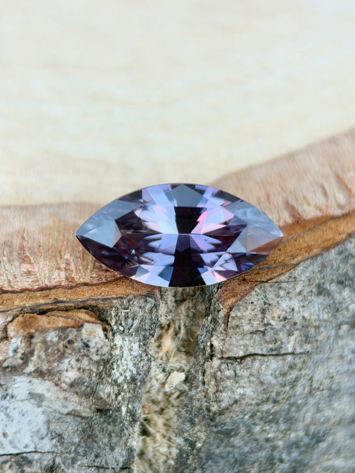 A marquise-cut purple sapphire with iridescent lavender and blue reflections, resting on a textured piece of wood against a light beige background.