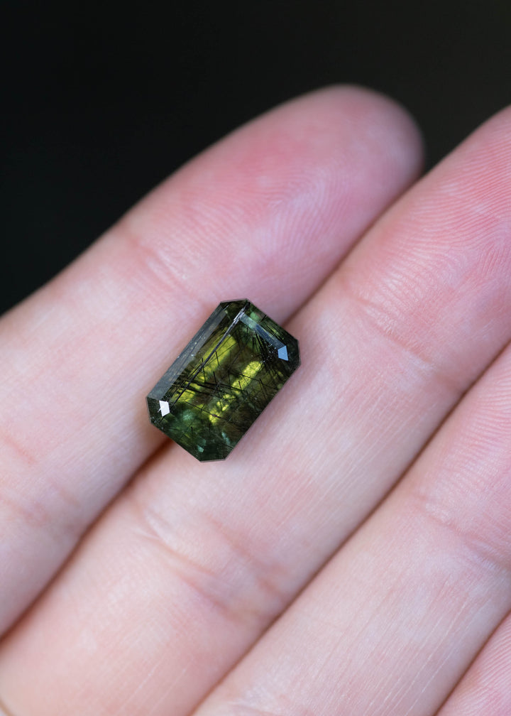 Natural untreated 6.00 carat deep forest green peridot from Skardu, Pakistan shown on fingertips for scale, displaying its elongated step mix cut and layered forest green tones.