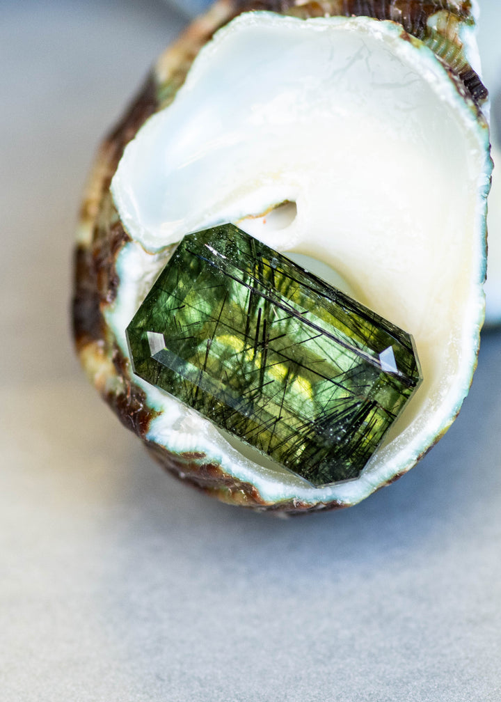 Natural untreated 6.00 carat deep forest green peridot from Skardu, Pakistan displayed in a shell, highlighting its step mix cut and dramatic natural needle inclusions.