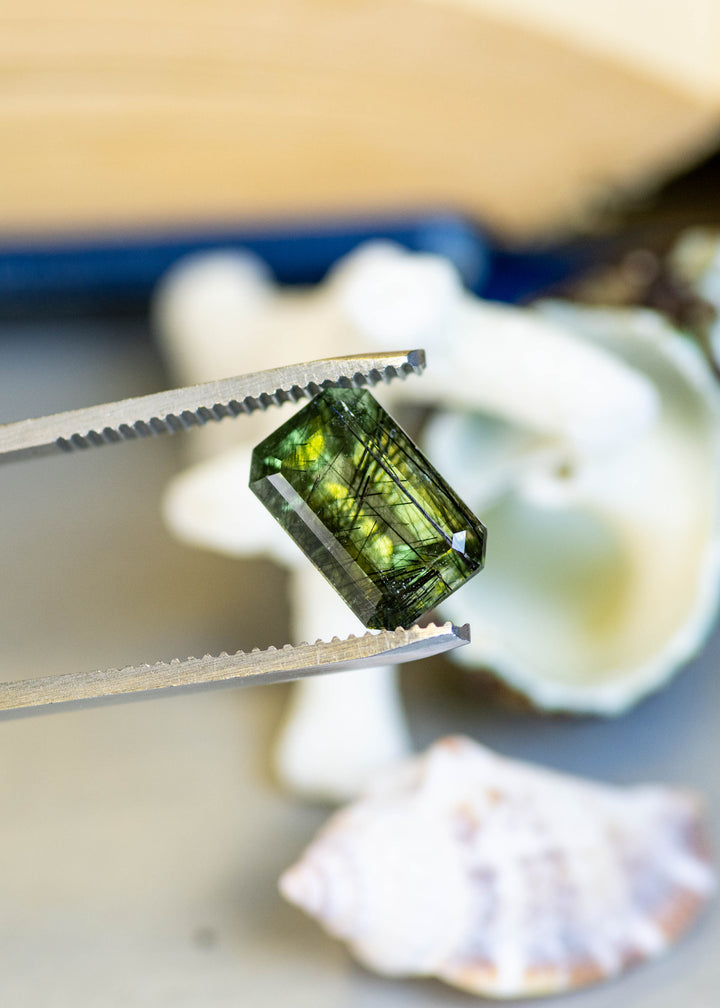 Close-up of a natural 6.00 carat deep forest green peridot from Pakistan held in tweezers, showcasing its elongated step mix cut and intricate crystal inclusions.