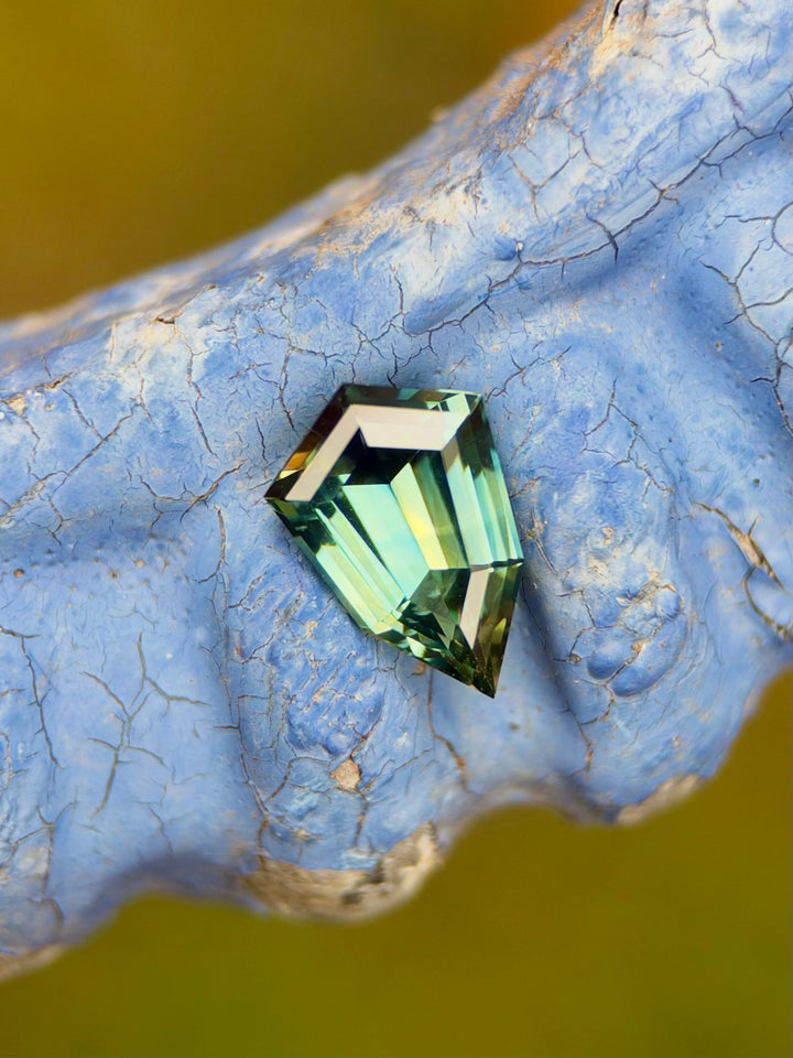 A faceted green sapphire with a geometric cut, displayed on a textured blue surface with a blurred olive-green background.