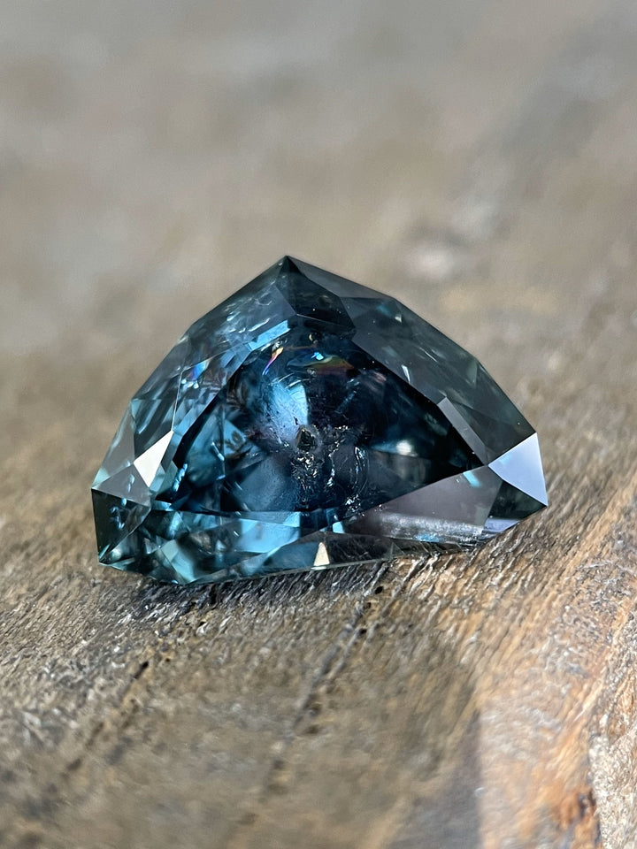 A 7.06-carat sapphire featuring a modified shield brilliant cut, with bluish-green transitioning to gray-purple tones, laid on a wooden surface.
