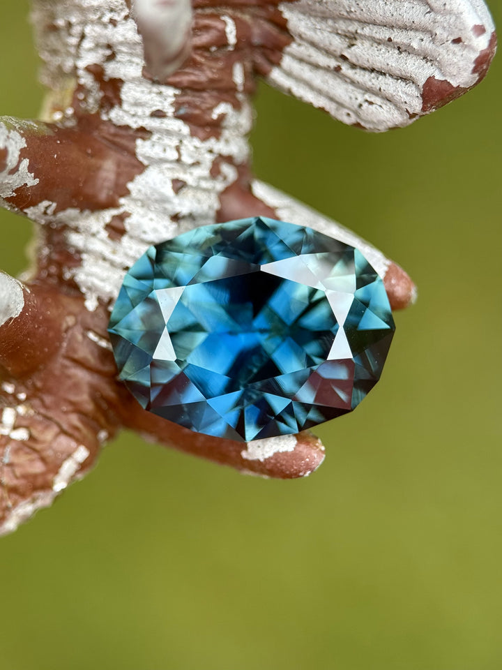 Faceted oval teal-blue sapphire, 7.75 carats, displayed on a rustic painted metal object with rich color zoning and sparkle.