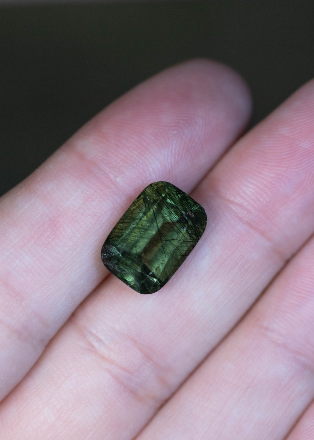 Natural untreated 8.25 carat deep forest green peridot from Skardu, Pakistan displayed on fingertips for scale, featuring an elongated cushion mix cut and layered forest green tones.
