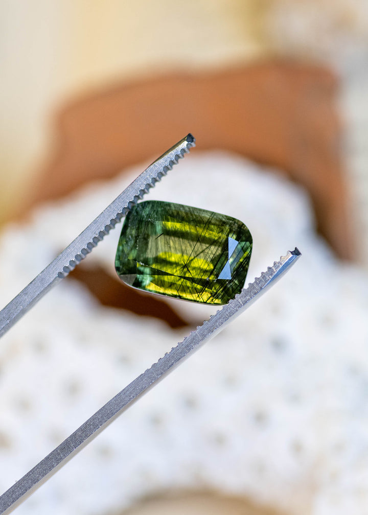 Close-up of a natural 8.25 carat deep forest green peridot from Pakistan held in tweezers, highlighting its elongated cushion mix cut, olive-green brilliance, and natural inclusions.