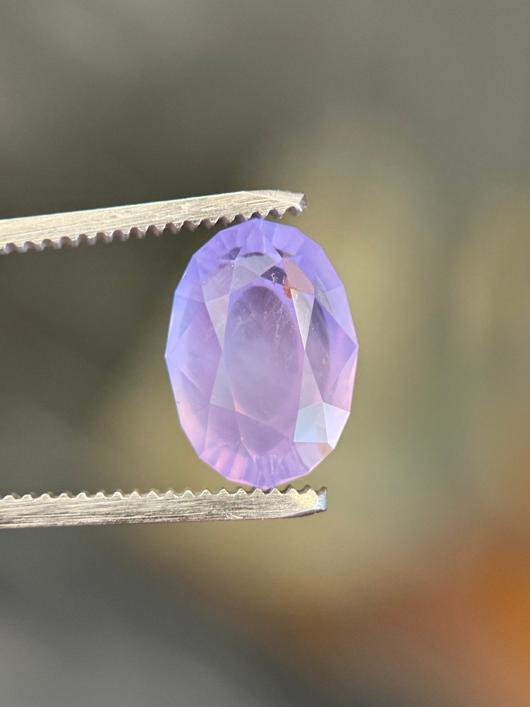 8.19 carat sapphire held in tweezers showing soft lavender violet tone and open culet facet.