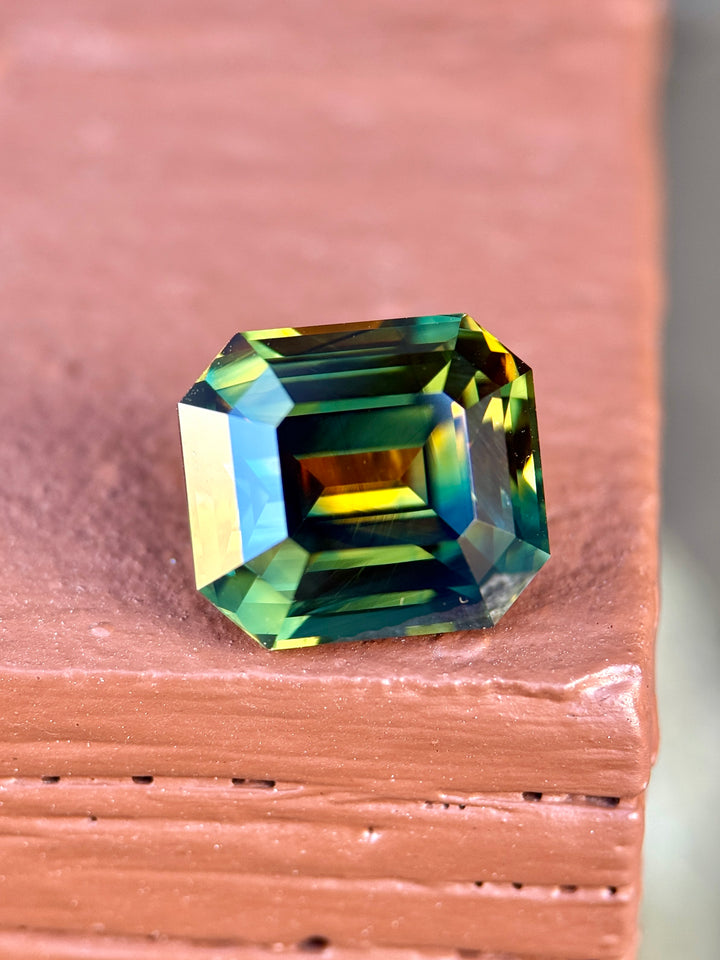 Large 9.14 carat emerald cut Nigerian parti sapphire resting on stone surface, vivid orange yellow blue green zoning with exceptional brilliance and depth