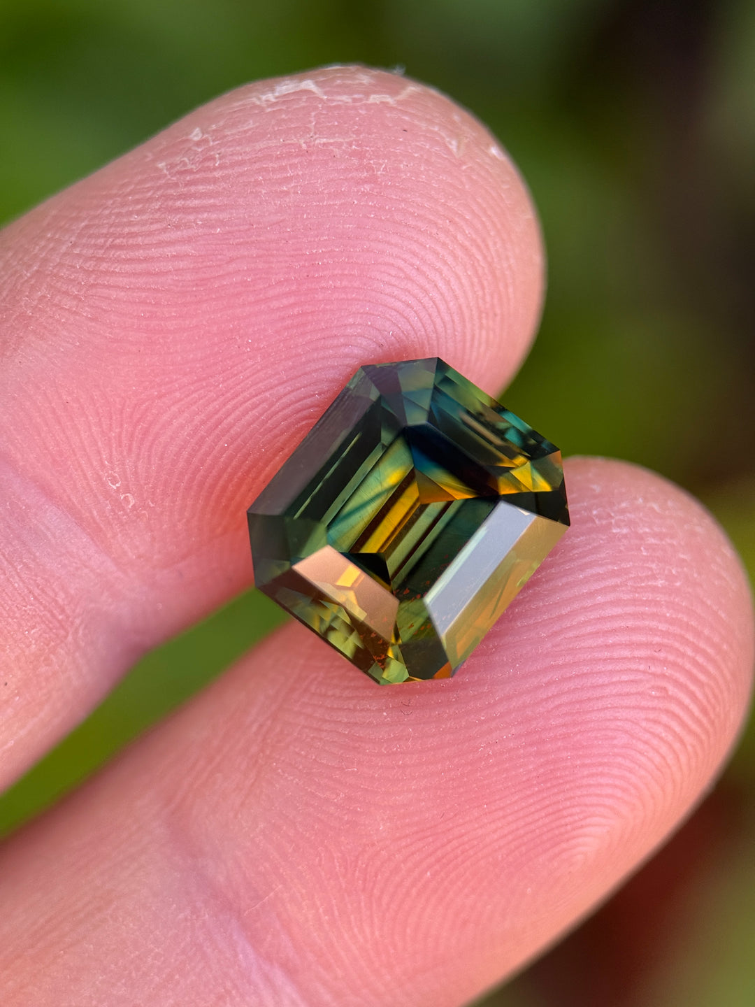 9.14 carat Nigerian parti sapphire held between fingers, precision emerald cut showing orange, yellow, blue, and green color zoning from Gombe, Nigeria
