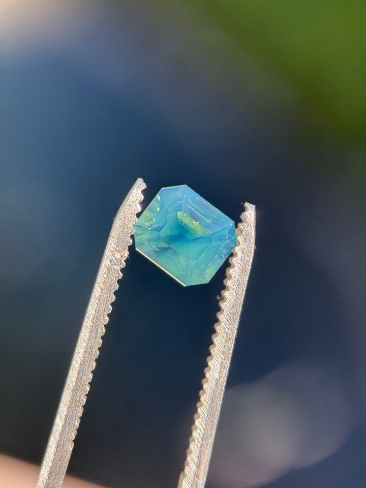 Teal opalescent sapphire held in gem tweezers under direct sunlight showing natural opalescence and vibrant blue/green hues. 