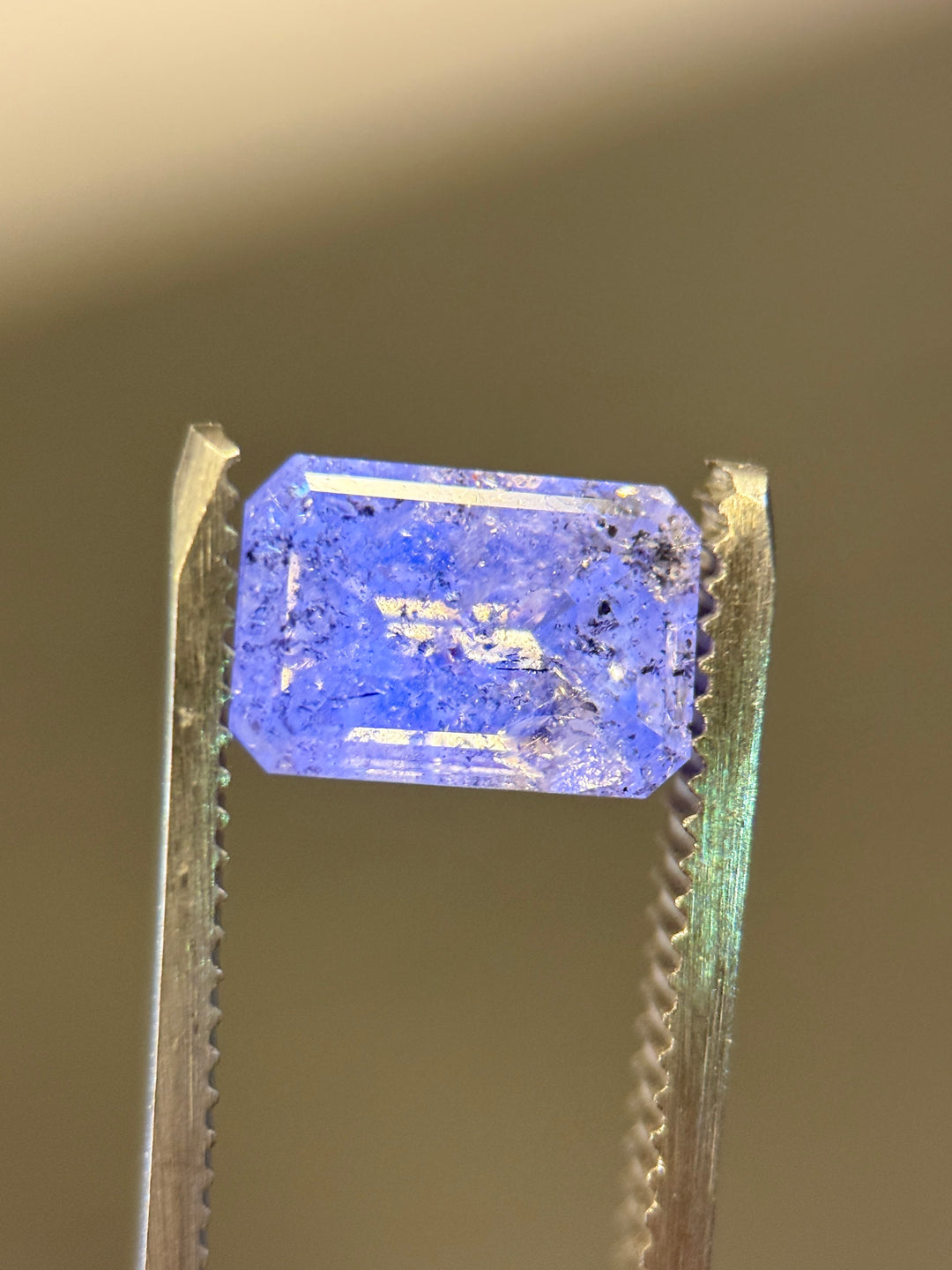 A rectangular emerald-cut blue gemstone held in forceps against a warm beige background.