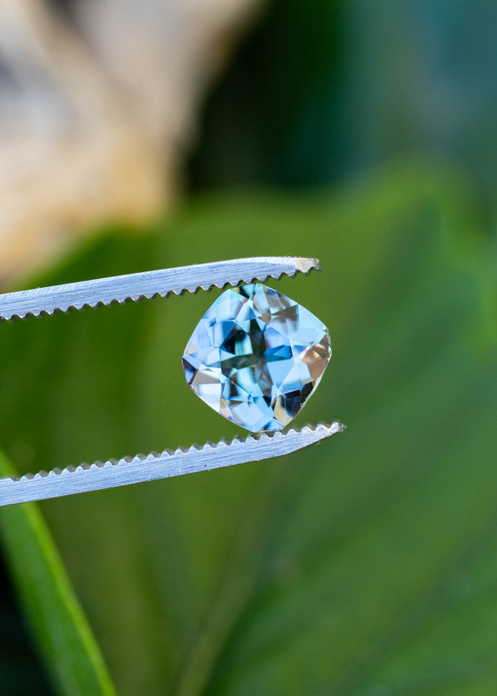 A custom square radiant cut Tanzanite held in jeweler's tweezers, showcasing bold geometric faceting and dramatic pleochroic shifts between teal, blue, and violet.