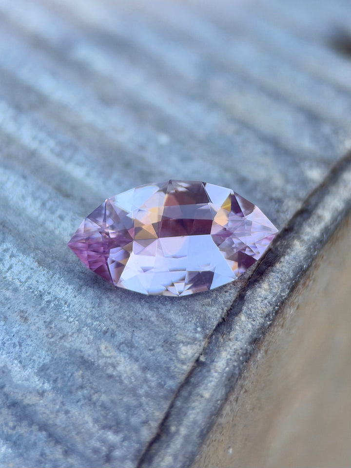 A marquise-cut pink sapphire with light lavender and purple tones, resting on a textured gray wooden surface.