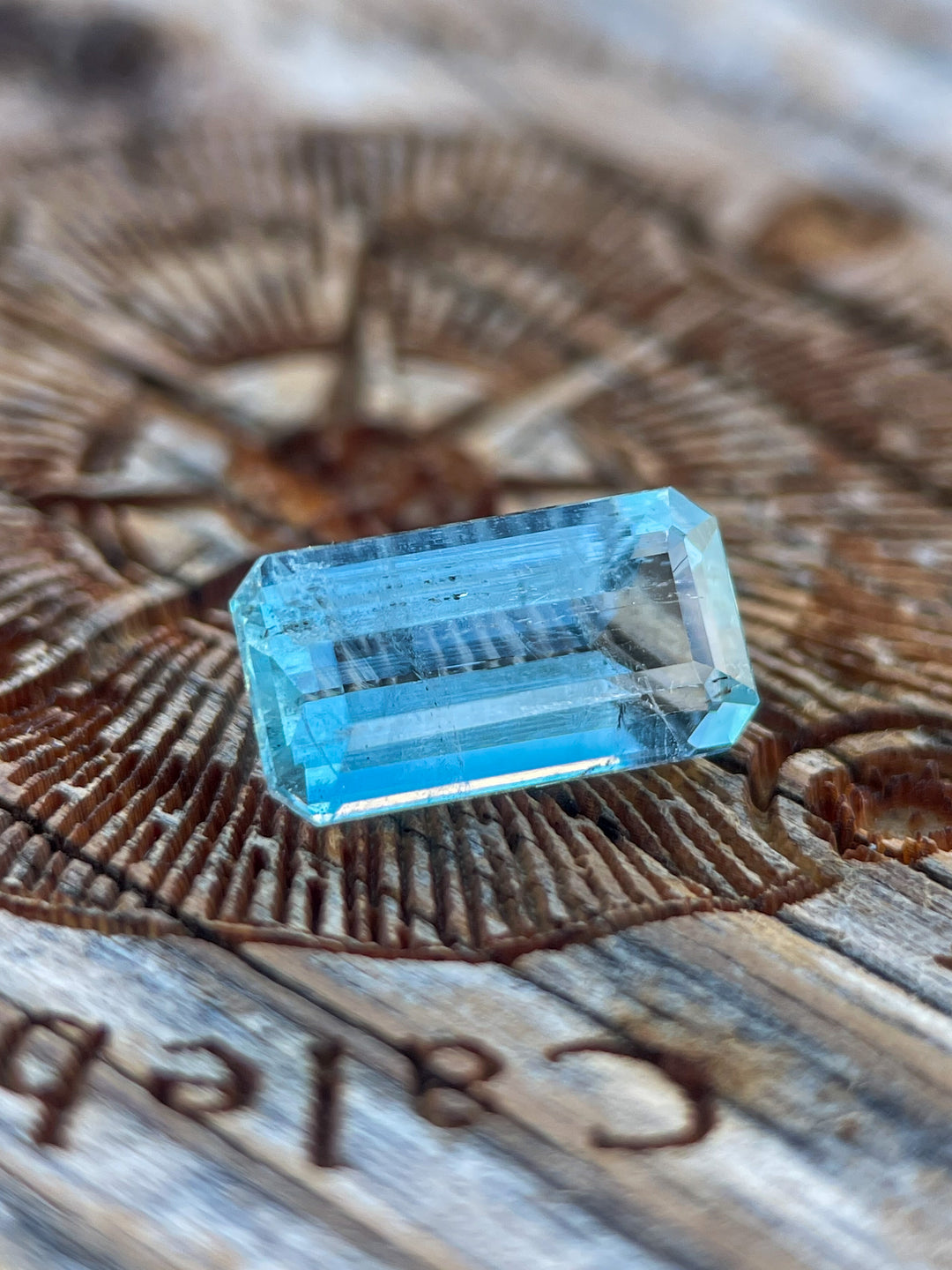 A mint blue emerald-cut aquamarine gemstone placed on a rustic wooden surface with intricate patterns, highlighting the gem’s unique color and craftsmanship.