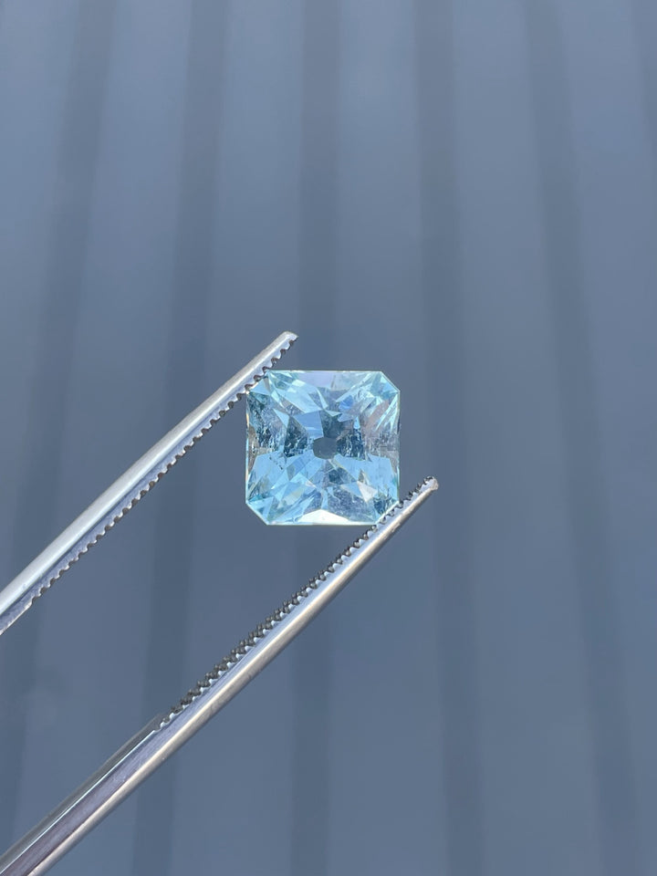 A dazzling aquamarine gemstone with blue and seafoam hues, weighing 2.87 carats, held by tweezers to showcase its custom antique cushion cut.