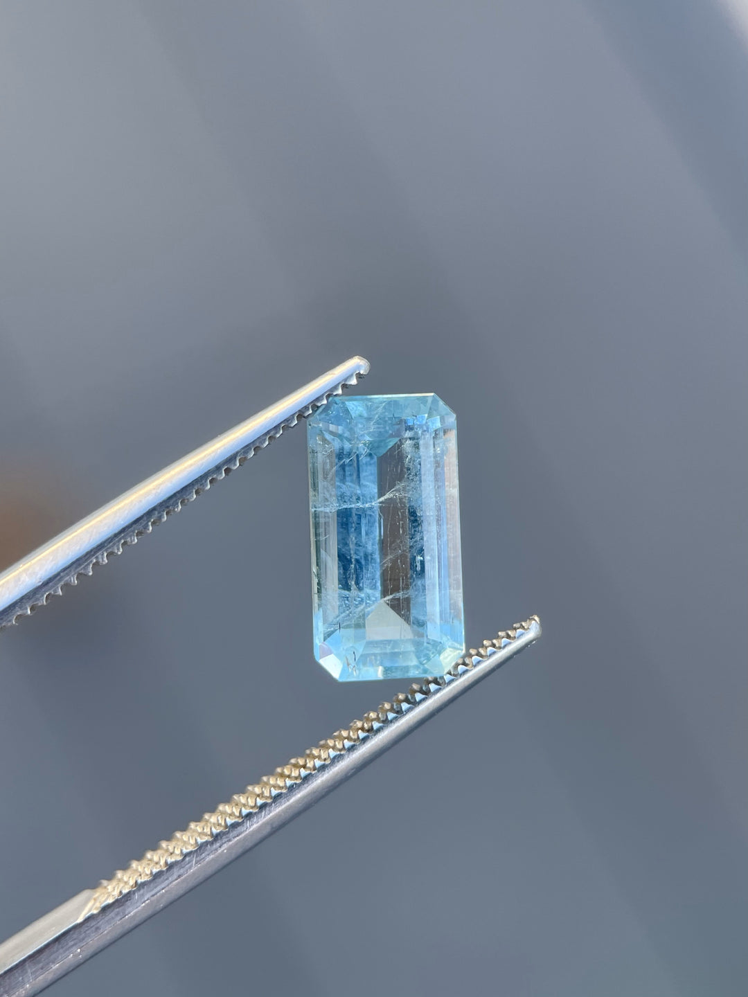 A mint blue emerald-cut aquamarine gemstone held in precision tweezers against a neutral gray background, emphasizing its elegant cut and clarity.