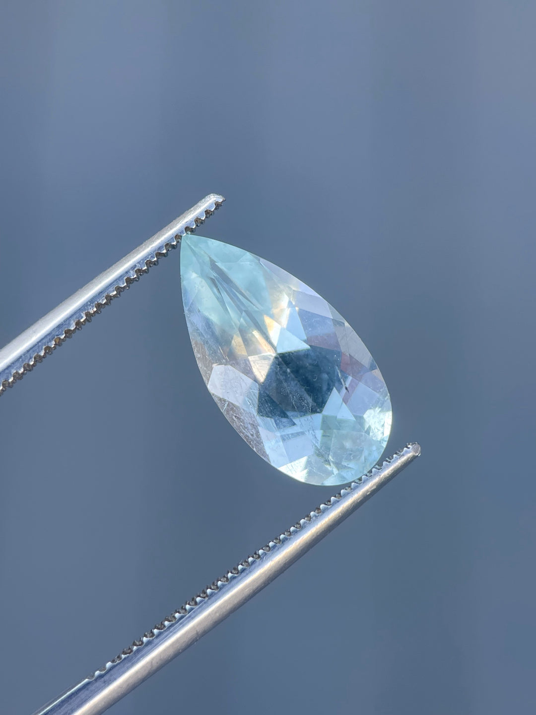 A 3.88-carat green mint blue aquamarine gemstone in a custom pear cut, held in tweezers against a muted background.