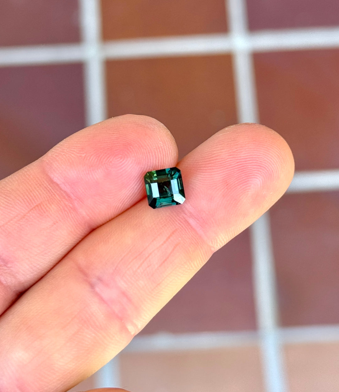 Asscher cut Kenyan sapphire resting on fingertips, showing deep lagoon teal color, step-cut brilliance, and architectural symmetry.