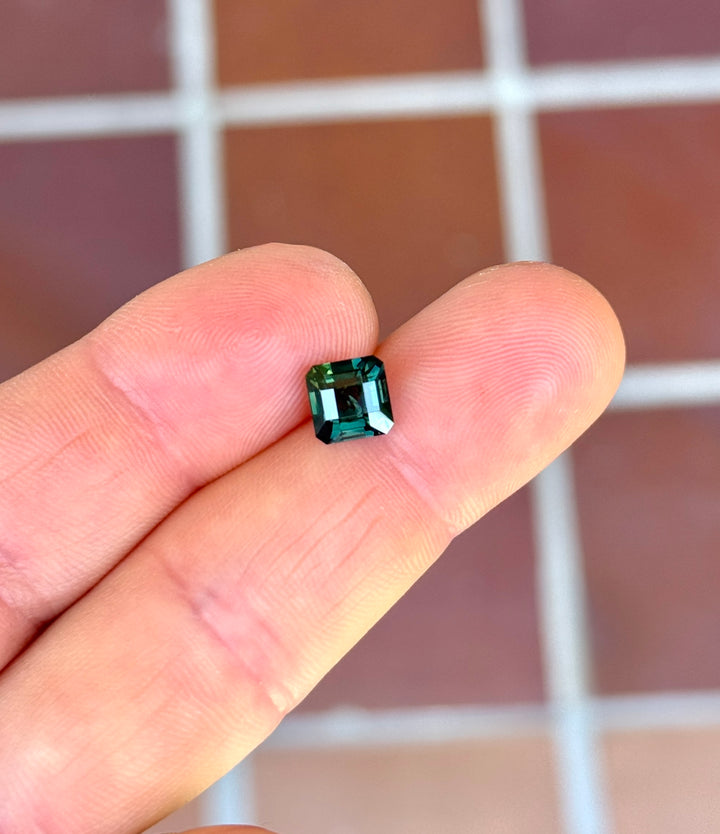 Asscher cut Kenyan sapphire resting on fingertips, showing deep lagoon teal color, step-cut brilliance, and architectural symmetry.