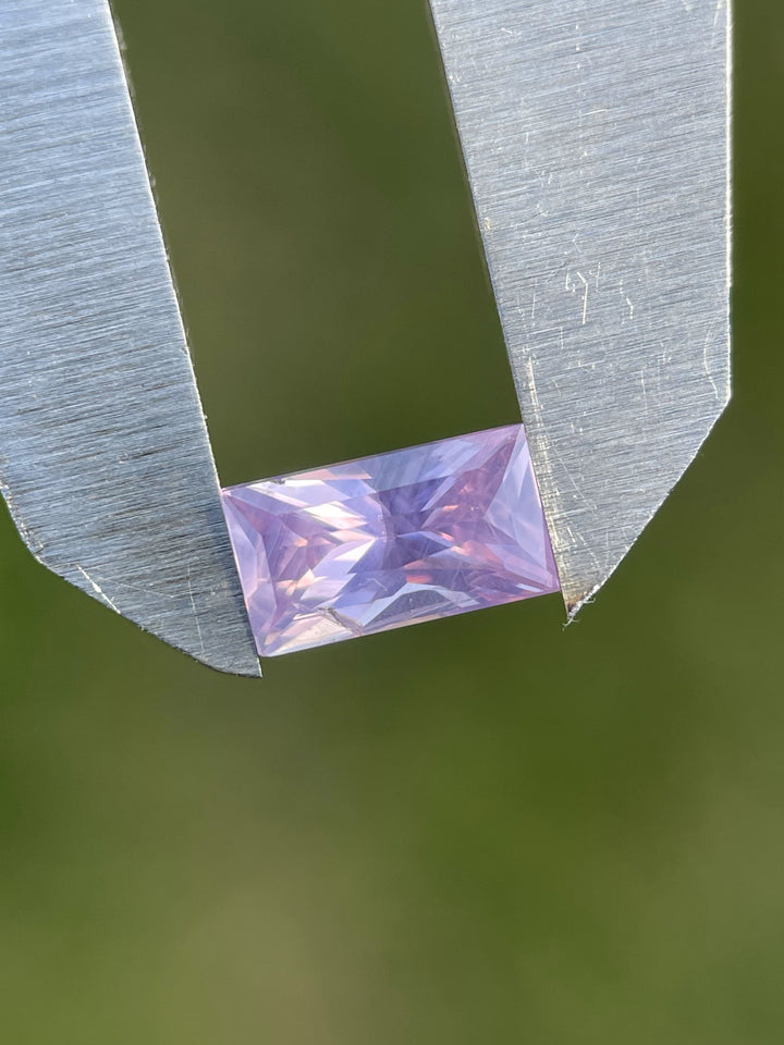 1.53ct pink and purple sapphire held in gem tweezers, highlighting its sharp baguette shape and pastel opalescence against a soft green blurred background.