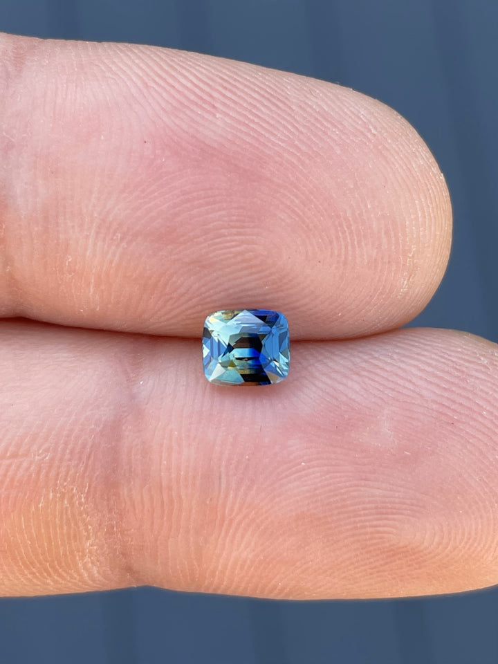 A stunning 1.02-carat bi-color sapphire featuring a mix of blue and yellow hues, displayed on a fingertip to showcase its cushion-brilliant cut and size.