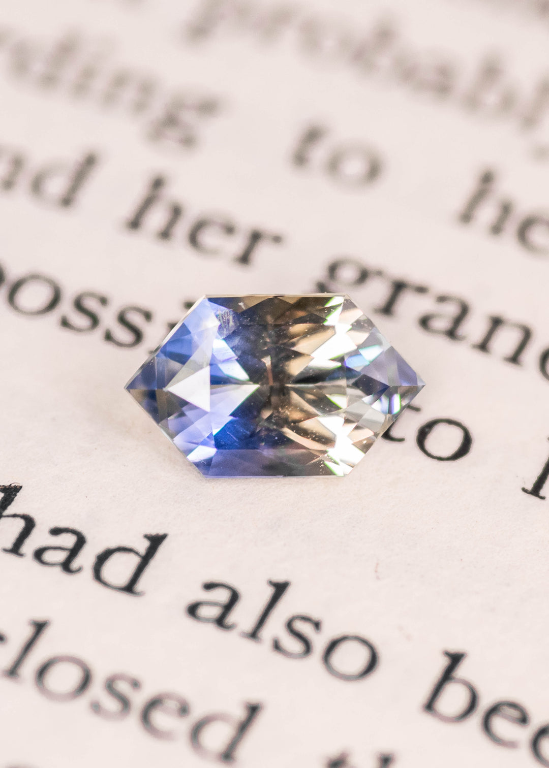 Bi-color parti sapphire with blue and yellow color zoning shown face-up on printed text, highlighting the elongated hexagonal mix cut, natural brilliance, and 1.57 carat size from Ratnapura, Sri Lanka.