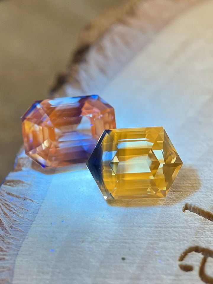 Close-up of two bi-color sapphires on a wood surface, showcasing radiant blue, yellow, and orange tones in hexagonal step cuts, with glowing saturation.
