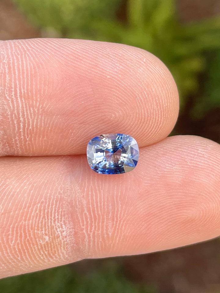A bi-color sapphire in mix brilliant cushion cut resting on a fingertip. Displays mauve and blue tones with a 2.28-carat weight. Heated and sourced from Sri Lanka.