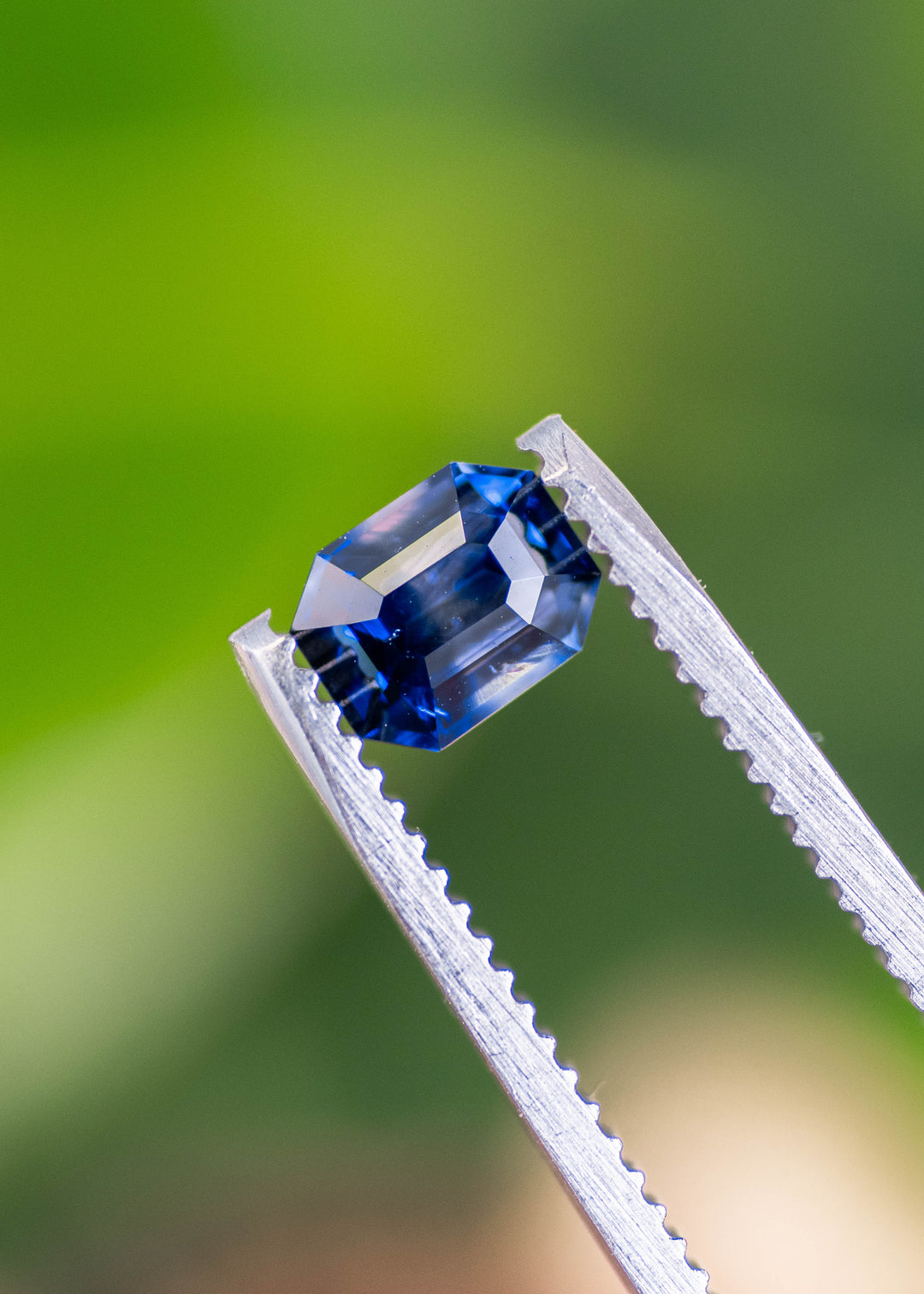 Loose blue bi-color sapphire from Sri Lanka held in tweezers highlighting the emerald cut shape and natural blue and colorless tones in the gemstone.