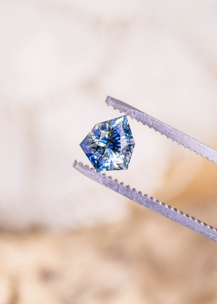 Loose blue and colorless sapphire from Madagascar held in tweezers displaying the geometric concave shield cut and the contrast between the blue and clear zones.