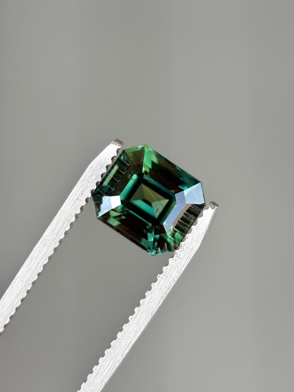 Loose blue green sapphire from Diego Madagascar held in tweezers highlighting the emerald step cut and rich teal color.
