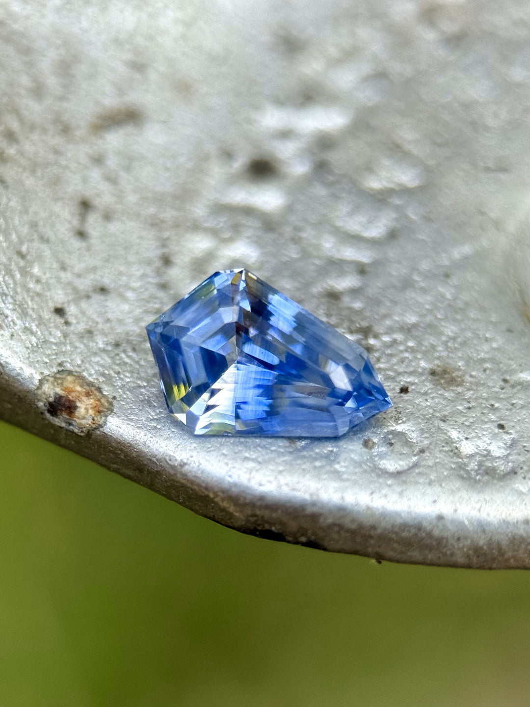 Close-up of a heated Parti sapphire with step-cut shield shape and strong color zoning, placed on a rustic metal surface.