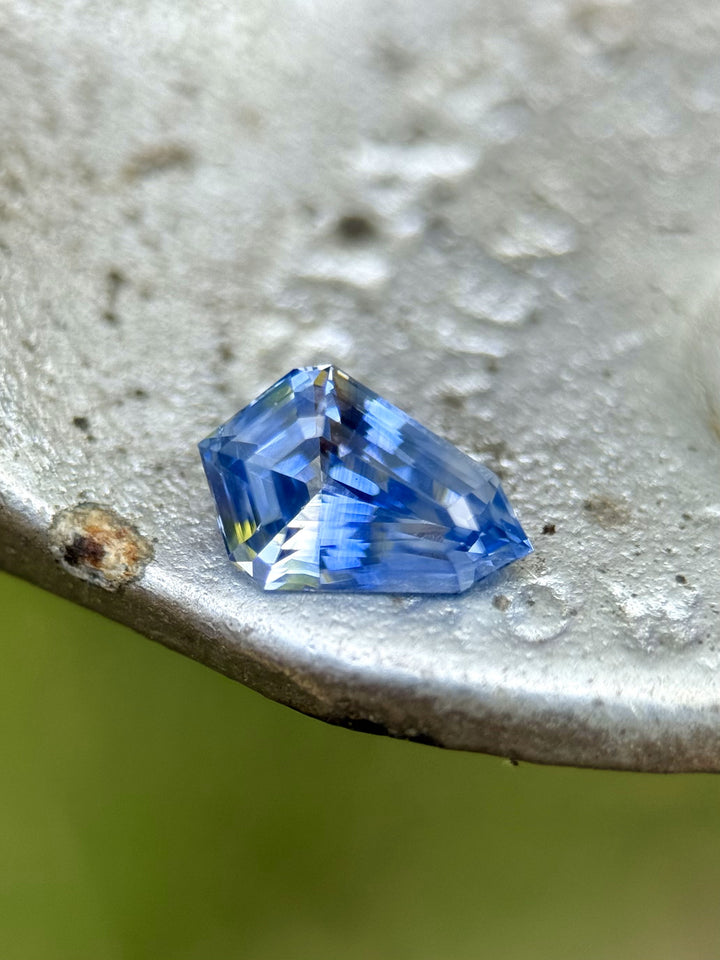 Close-up of a heated Parti sapphire with step-cut shield shape and strong color zoning, placed on a rustic metal surface.