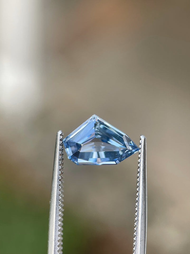 A stunning blue sapphire weighing 2.23 carats, cut in a free-form geometric shape, showcased between tweezers against a natural background. Origin: Sri Lanka, Untreated.