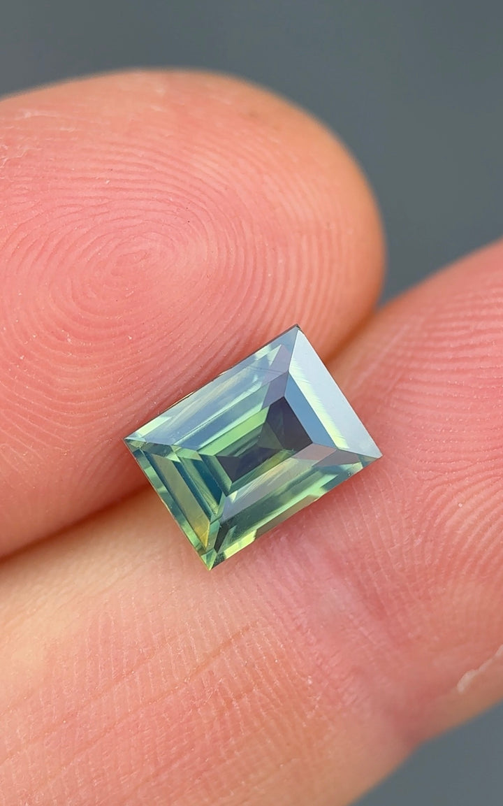 Blue green opalescent baguette sapphire held between fingers against a neutral background