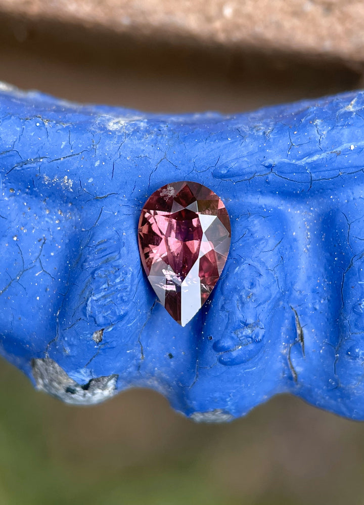 0.88 carat blush red sapphire placed on a textured blue surface, emphasizing its untreated beauty and vibrant pear-shaped sparkle.