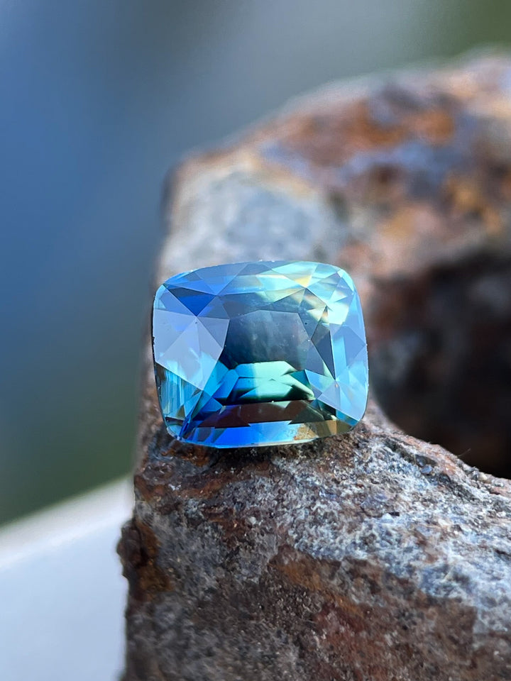 Close-up of a 1.02-carat cushion-cut bi-color sapphire from Madagascar, highlighting its rich blue and golden tones on a textured rock surface.