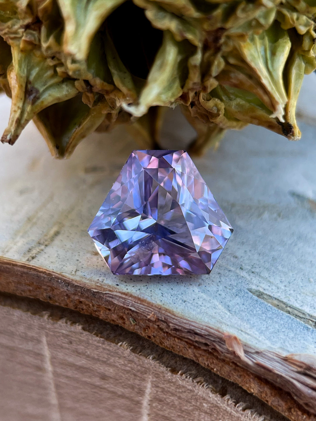 A loose 1.56-carat lavender sapphire resting on a natural surface, displaying its unique custom brilliant triangle cut with a mix of purple, pink, and lavender tones.