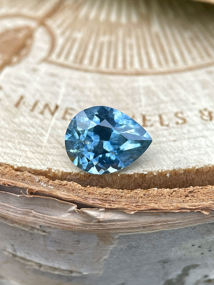 Close-up of a 1.70 carat sapphire from Montana resting on a wooden surface, featuring a rich blue color and custom brilliant pear faceting.