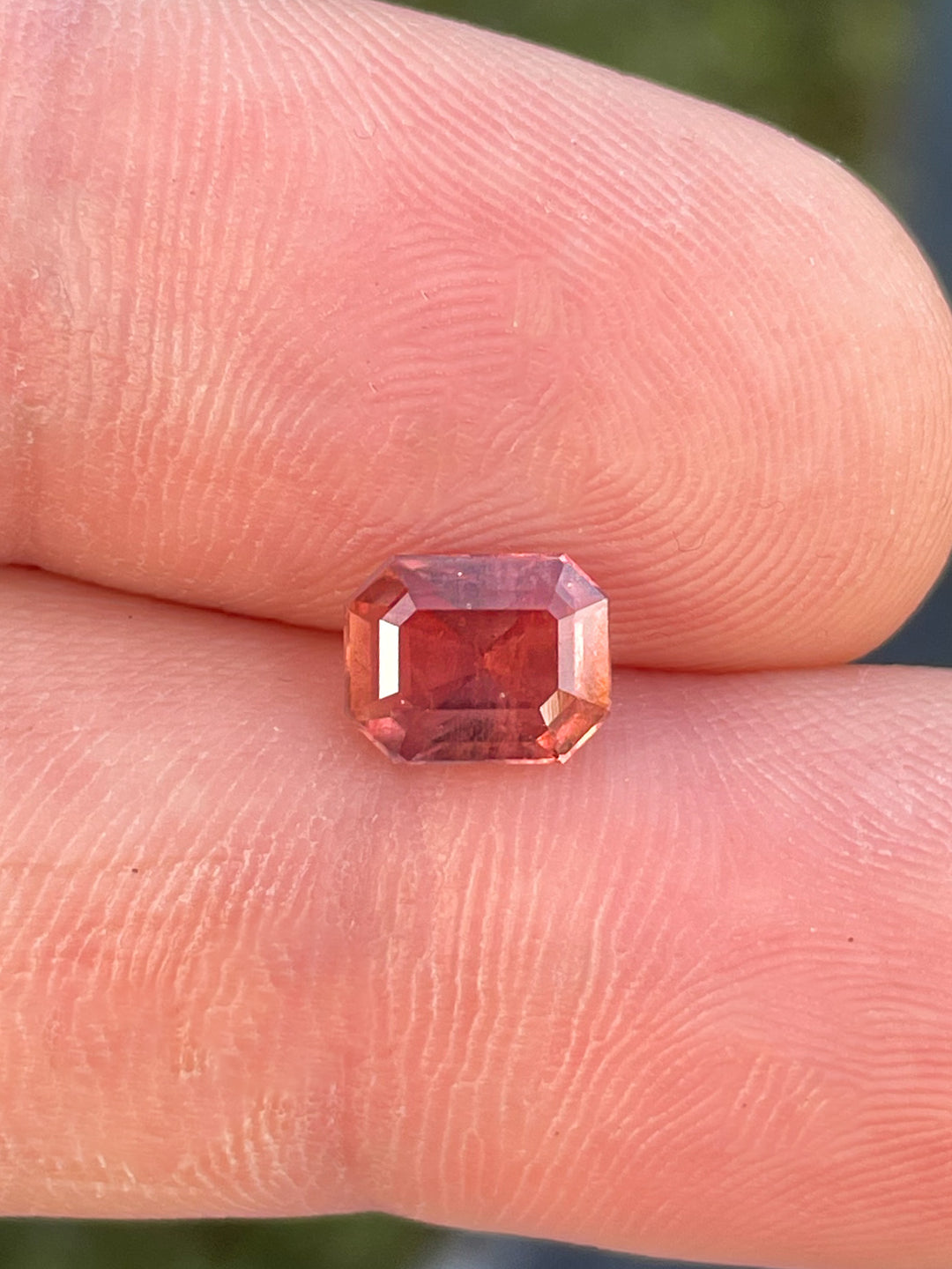 A close-up of a Cabernet-colored sapphire resting on a person's fingertip, showcasing its modified emerald cut and rich color.